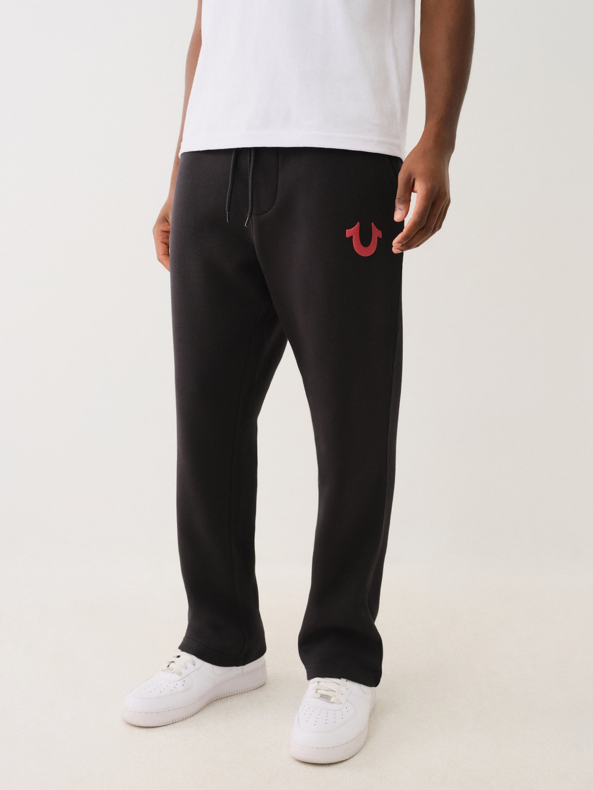Pantalones de sudor negros con logo rojo, estilo casual, ideales para hombre con zapatillas blancas.